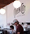 Barista in a coffee shop with 'Queen City Collective Coffee' branding on the wall.